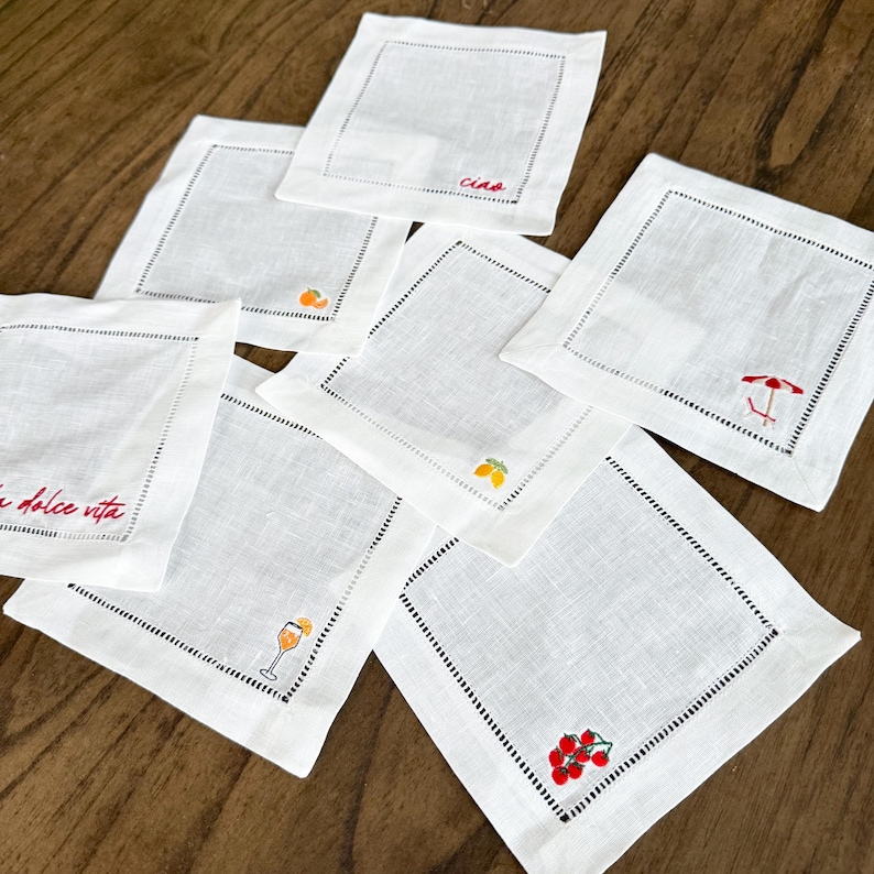 May include: White linen coasters with embroidered details. Some coasters feature the text "Ciao" and "La dolce vita" in red. Other coasters have embroidered images of a cocktail, fruit, and a beach umbrella.