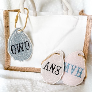 May include: A white canvas tote bag with a burlap trim and a rope handle. Attached are three oyster-shaped keychains with embroidered initials in black and blue. The keychains are attached with gold-colored clasps.