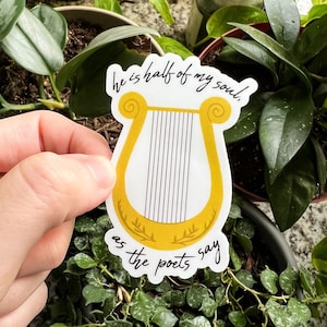 Pode incluir: Um autocolante branco com uma ilustra&ccedil;&atilde;o de lira dourada e o texto "he is half of my soul, as the poets say" em escrita preta. O autocolante &eacute; segurado em frente a um fundo de plantas verdes.