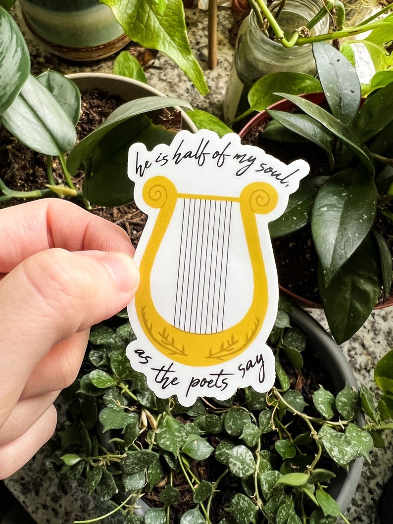 Pode incluir: Um autocolante branco com uma ilustra&ccedil;&atilde;o de lira dourada e o texto "he is half of my soul, as the poets say" em escrita preta. O autocolante &eacute; segurado em frente a um fundo de plantas verdes.