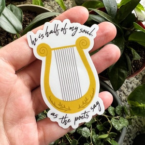Pode incluir: Um autocolante com um desenho de harpa dourada e cordas pretas, com a cita&ccedil;&atilde;o "he is half of my soul as the poets say" em escrita preta. O autocolante &eacute; segurado em uma m&atilde;o, com plantas verdes no fundo.
