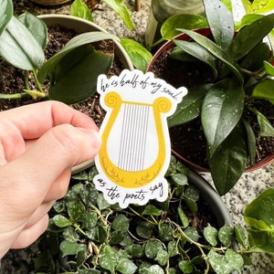 Pode incluir: Um autocolante com uma lira amarela com cordas pretas e o texto "he is half of my soul, as the poets say" em escrita preta. O autocolante &eacute; segurado em frente de v&aacute;rias plantas em vasos.