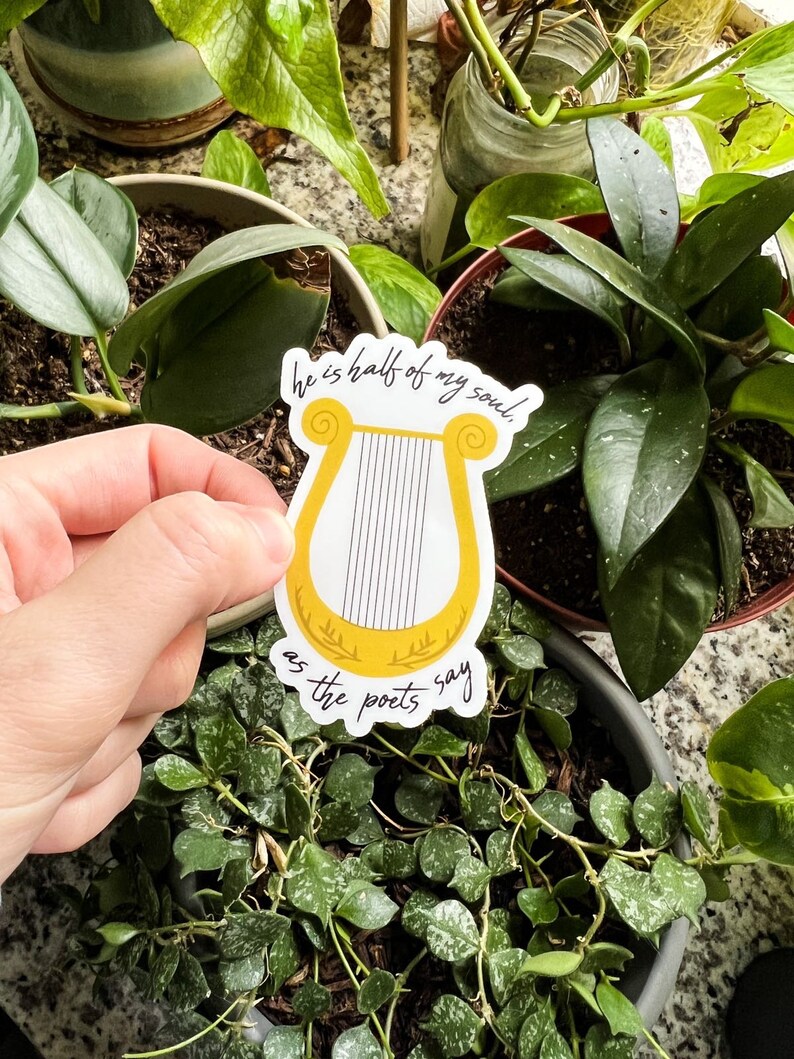 Pode incluir: Um autocolante com uma lira amarela com cordas pretas e o texto "he is half of my soul, as the poets say" em escrita preta. O autocolante &eacute; segurado em frente de v&aacute;rias plantas em vasos.