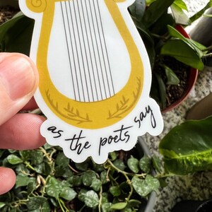 Pode incluir: Um autocolante com uma lira dourada com cordas pretas. O autocolante tem o texto "he is half of my soul" na parte superior e "as the poets say" na inferior. O autocolante est&aacute; colocado sobre um fundo de plantas verdes.