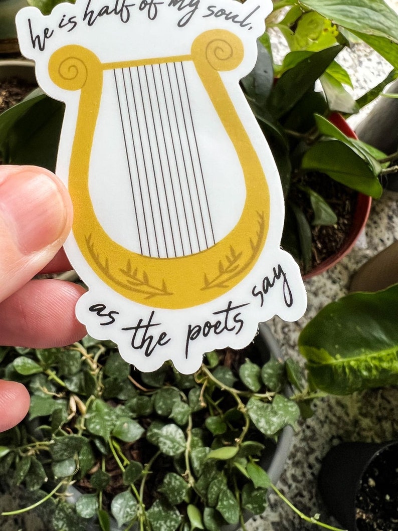 Pode incluir: Um autocolante com uma lira dourada com cordas pretas. O autocolante tem o texto "he is half of my soul" na parte superior e "as the poets say" na inferior. O autocolante est&aacute; colocado sobre um fundo de plantas verdes.