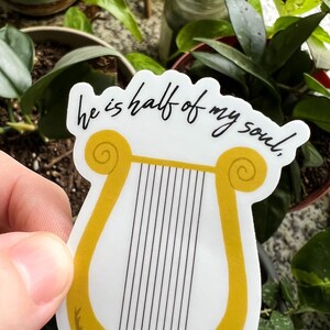 Pode incluir: Um autocolante branco com uma lira dourada e cordas pretas, com o texto "he is half of my soul". O autocolante tem um desenho de louro na base. O fundo mostra plantas verdes. O autocolante &eacute; segurado em frente das plantas.
