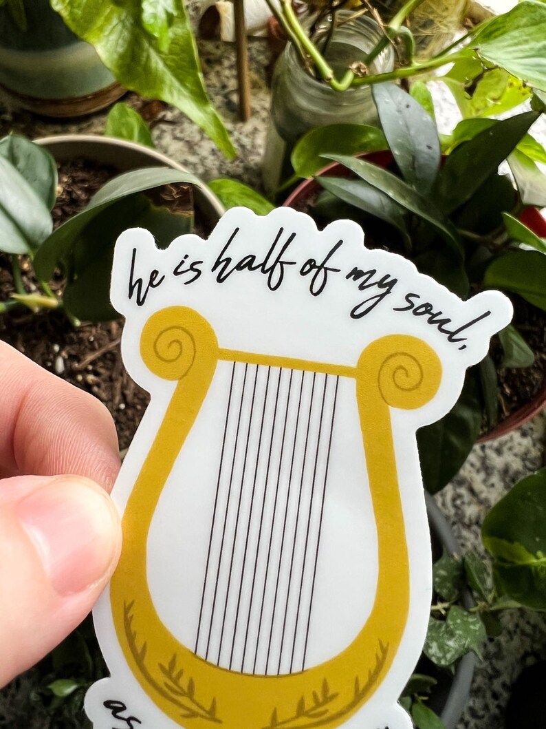 Pode incluir: Um autocolante branco com uma lira dourada e cordas pretas, com o texto "he is half of my soul". O autocolante tem um desenho de louro na base. O fundo mostra plantas verdes. O autocolante &eacute; segurado em frente das plantas.
