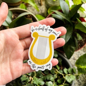 Pode incluir: Um autocolante com uma lira dourada com o texto "he is half of my soul, as the poets say" em escrita preta. O autocolante &eacute; segurado por uma m&atilde;o, com plantas verdes no fundo.
