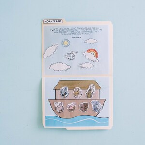 May include: A colorful illustration of Noah's Ark with various animal pairs. The top section features clouds, a rainbow, and the sun. The text "NOAH'S ARK" is at the top. The bottom section shows the ark with animals. The background is light blue.