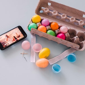 May include: A cardboard egg carton filled with colourful plastic Easter eggs. There are also several empty plastic eggs, a small wooden cross, a pin, and a smartphone displaying a photo of two people on a beach.