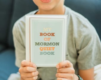 Peut inclure: Une personne tient un livre intitulé "MY BOOK OF MORMON QUIET BOOK". Le livre a un fond bleu clair avec du texte orange, vert et gris. La personne porte un t-shirt gris et un pantalon marron.