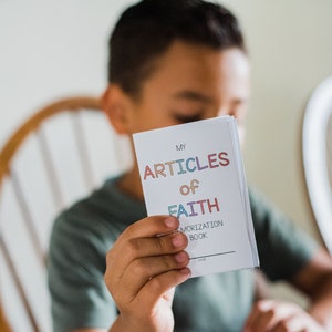 May include: A child holds a white booklet titled "My Articles of Faith Memorization Book." The title text is in colorful letters. The child is wearing a green shirt and sitting in front of a wooden chair.