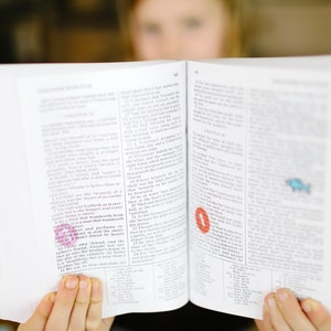 Op de afbeelding: Een jong persoon leest een wit boek met een rode en roze markeerstift. Het boek is open op een pagina met tekst en een kleine illustratie van een vis.