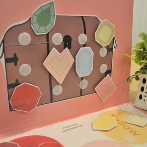 May include: A pop-up book with a brown treasure chest illustration and colorful gem stickers. The gems are various shapes and colors, including green, yellow, and pink. The book is open on a pink background.