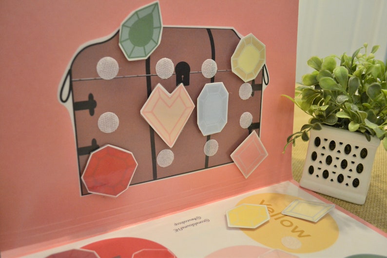May include: A pop-up book with a brown treasure chest illustration and colorful gem stickers. The gems are various shapes and colors, including green, yellow, and pink. The book is open on a pink background.