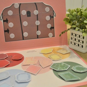 May include: An open book with a pink cover and colorful shapes. The left page has red, pink, and blue shapes. The right page has yellow and green shapes. A brown treasure chest illustration is on the left page.