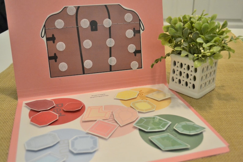 May include: An open book with a pink cover and colorful shapes. The left page has red, pink, and blue shapes. The right page has yellow and green shapes. A brown treasure chest illustration is on the left page.