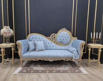 May include: A light blue chaise lounge with gold trim and tufted back. Two matching pillows and a decorative throw are on the chaise. The room includes two small side tables, a patterned rug, and a floral lamp.