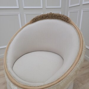 Pu&ograve; includere: Una poltrona imbottita bianca con schienale arrotondato di forma ovale e una bordatura decorativa attorno alla parte superiore e inferiore. La poltrona &egrave; in tessuto beige chiaro e ha una struttura in legno.