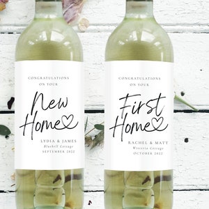 Puede incluir: Dos botellas de vino blanco con etiquetas que dicen "Felicidades por tu nueva casa" y "Felicidades por tu primera casa". Las etiquetas tienen una fuente manuscrita y un pequeño símbolo de corazón. Las etiquetas también incluyen los nombres de las parejas y las fechas de compra de su nueva casa.