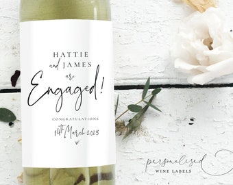 Può includere: Una bottiglia di vino bianco con un'etichetta personalizzata che dice "Hattie e James sono fidanzati!" con la data "14 marzo 2023". L'etichetta è bianca con testo nero e un simbolo di cuore. L'etichetta include anche il testo "Congratulazioni".