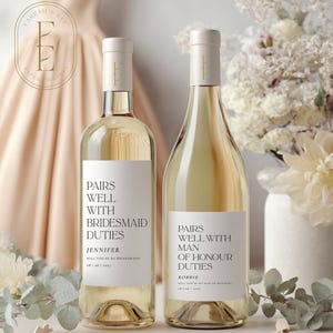 Op de afbeelding: Twee flessen witte wijn met aangepaste etiketten. De etiketten luiden "Pairs Well With Bridesmaid Duties" en "Pairs Well With Man of Honour Duties". De flessen worden gepresenteerd met bloemstukken.