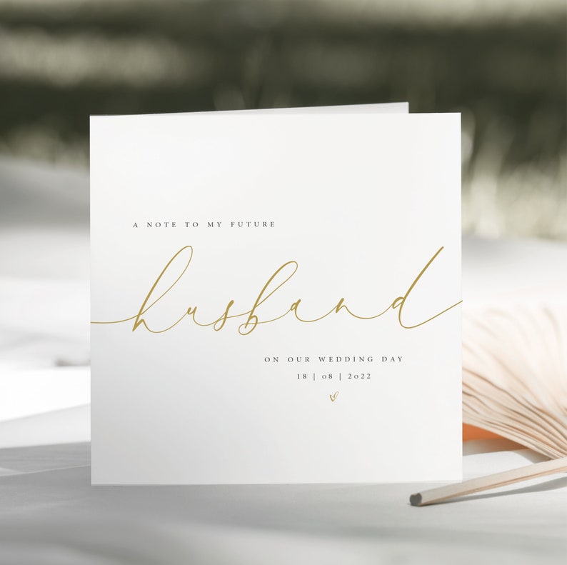 May include: A white card with gold lettering that reads "A note to my future husband on our wedding day 18 | 08 | 2022".