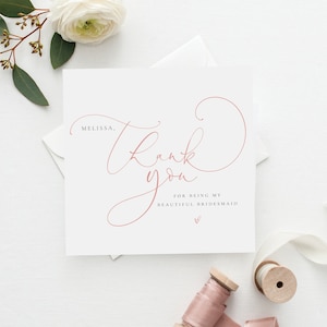 Może przedstawiać: Kwadratowa, kremowa karta z napisem "Thank you" w płynnym, różowym skrypcie. Na karcie widnieje napis "Melissa, Thank you for being my beautiful bridesmaid" delikatną czcionką. Na dole małe serce.