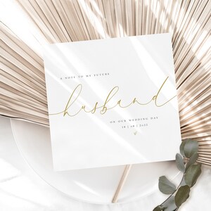 May include: A white card with gold lettering that reads "A note to my future husband on our wedding day 18 | 08 | 2022". The card is sitting on a white plate with a beige fan-shaped background.