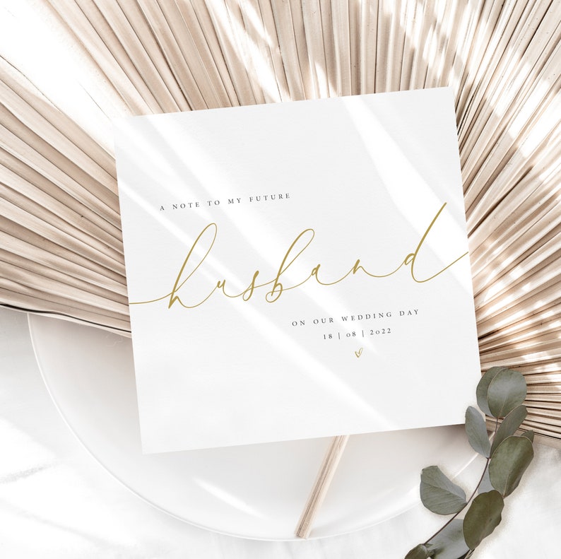 May include: A white card with gold lettering that reads "A note to my future husband on our wedding day 18 | 08 | 2022". The card is sitting on a white plate with a beige fan-shaped background.