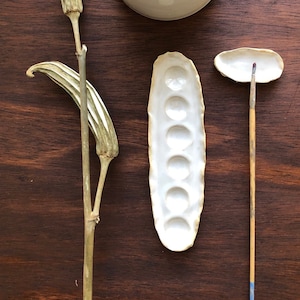 May include: A collection of ceramic art supplies, including a small bowl, a paint palette with seven wells, and a small dish with a wooden handle. A dried okra plant is also included. All items are white and arranged on a dark wood surface.