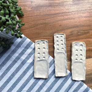 Puede incluir: Tres platos rectangulares de cerámica blanquecina con superficie texturizada e indentaciones circulares. Los platos están dispuestos sobre un paño a rayas azules y blancas, con una superficie de madera en el fondo. Una planta en maceta es visible en la esquina superior izquierda.