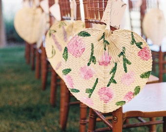 May include: A row of wooden chairs with hand-painted fans tied to the back. The fans are white with pink flowers and green leaves.