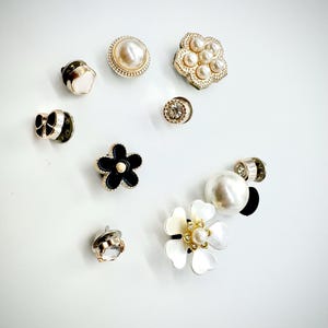 May include: An assortment of decorative buttons in various designs. The buttons include pearl, floral, and geometric shapes, with gold and black accents. Some buttons feature faux pearls and clear crystals. The buttons are arranged on a white surface.