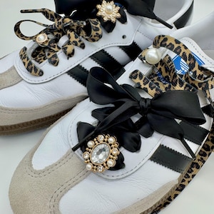 May include: A pair of white sneakers with black stripes and leopard print shoelaces. The laces are tied with black satin ribbon bows and embellished with jeweled brooches.