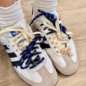 Puede incluir: Zapatillas blancas y grises con cordones azul marino y adornos decorativos. Las zapatillas tienen el logo de Adidas y tres rayas negras. La puntera y el talón son de ante gris. Los cordones están adornados con perlas y detalles dorados.