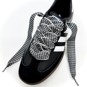 May include: Black and white Adidas Samba sneaker with white stripes and a brown sole. The shoe features black and white gingham patterned laces, creating a stylish contrast. The sneaker is set against a white background.