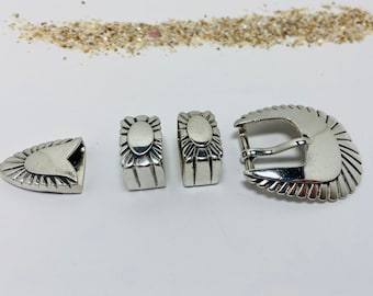 Puede incluir: Un conjunto de hebillas de cinturón de metal plateado, que incluye una hebilla, dos presillas y una punta. Las piezas tienen un diseño decorativo de rayos de sol y están dispuestas sobre una superficie blanca.