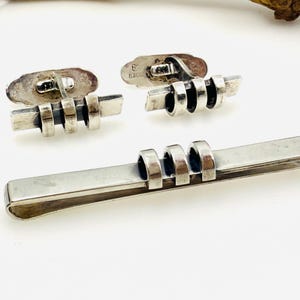 May include: A set of silver cufflinks and a tie bar with a geometric design. The cufflinks are rectangular in shape with three circular elements. The tie bar has a similar design with four circular elements.