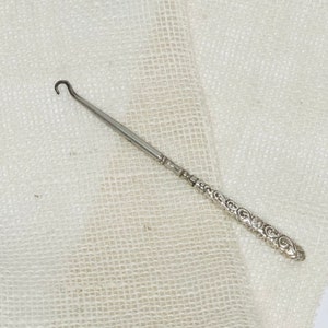 May include: A silver shoe horn with an ornate, floral design on the handle. The shoe horn is lying on a white, textured surface.
