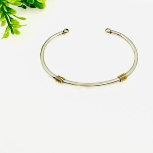 May include: A silver bangle bracelet with two gold accents. The bracelet is open and has a rounded shape.