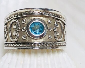 Puede incluir: Un anillo de plata con una piedra preciosa azul engastada en una banda decorativa. La banda presenta intrincados trabajos de volutas y un borde en forma de cuerda.