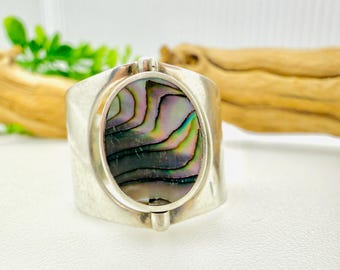 May include: A silver ring with an oval-shaped abalone shell inlay. The ring has a wide band with a textured finish.