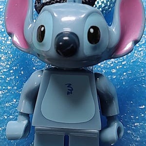 May include: A light blue Lego figure of the character Stitch from the Disney movie Lilo & Stitch. The figure has large pink ears and a black nose. The figure is attached to a blue and black woven cord.