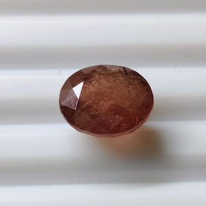 Puede incluir: Gema de corte ovalado con superficie facetada. La gema tiene un color marrón rojizo cálido con inclusiones internas. La piedra está sobre un fondo blanco, resaltando su forma y color. Adecuada para la fabricación de joyas.