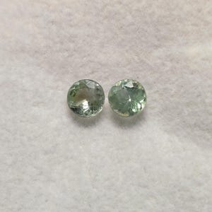 May include: Two round, faceted gemstones with a pale green hue. The gems are displayed on a white, textured surface. The gemstones have a brilliant cut, reflecting light.