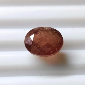 May include: An oval-shaped, faceted gemstone with a deep reddish-brown hue. The stone features a polished surface with multiple geometric facets that reflect light, showcasing internal textures and a translucent, warm-toned appearance throughout.