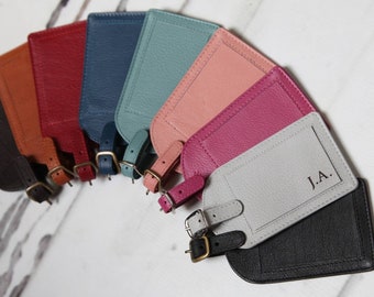 May include: A set of nine leather luggage tags in various colors, including brown, blue, teal, pink, magenta, gray, and black. Each tag has a metal buckle and a loop for attaching to luggage. The gray tag has the initials "J.A." embossed on it.