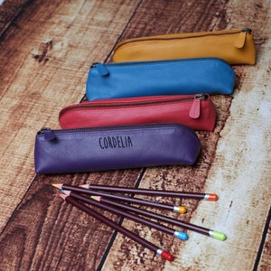 Op de afbeelding: Vier leren potloodkokers in verschillende kleuren: geel, blauw, rood en paars. De paarse koker heeft de naam "Cordelia" erop gegraveerd. Zes kleurpotloden liggen op een houten oppervlak.