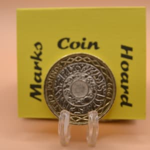 May include: A silver coin with a design of a circle with a smaller circle inside. The coin is on a yellow card with the text "Coin Hoard Marks" written on it.